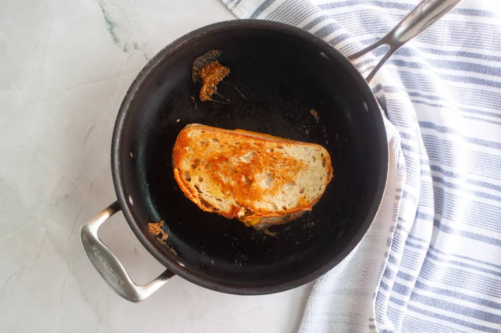 A grilled cheese sandwich being cooked in a black skillet on a marble countertop next to a white and blue striped towel.