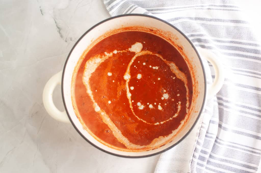 A white pot filled with red tomato soup topped with a swirl of cream, placed on a marble surface next to a striped kitchen towel.
