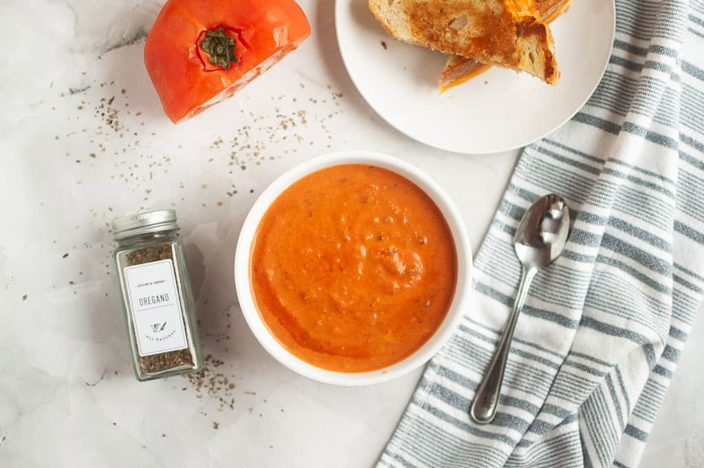 A bowl of tomato soup beside a sliced tomato, a jar of oregano, a spoon, a striped napkin, and a plate with a grilled cheese sandwich.