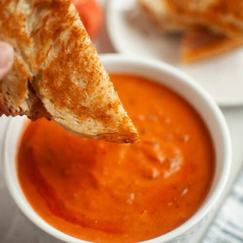 A grilled cheese sandwich is being dipped into a bowl of creamy tomato soup, with another sandwich half on a plate in the background.