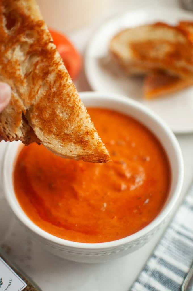 A grilled cheese sandwich is being dipped into a bowl of creamy tomato soup, with another sandwich half on a plate in the background.