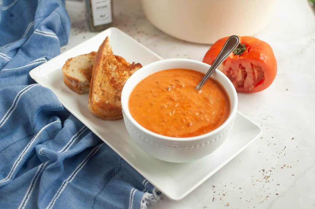 A bowl of tomato soup with a spoon, served on a rectangular plate with two slices of toasted bread and a halved tomato nearby.