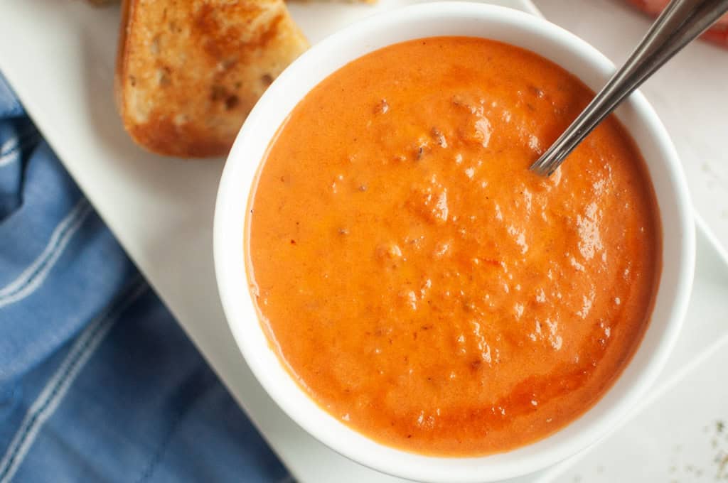 A bowl of creamy tomato soup with a spoon, served beside pieces of toasted bread on a white plate.