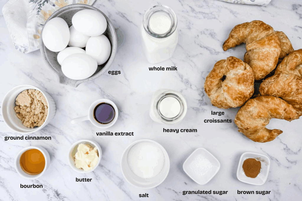 A tabletop displays labeled ingredients for baking: eggs, whole milk, croissants, heavy cream, brown sugar, ground cinnamon, bourbon, vanilla extract, butter, granulated sugar, and salt.