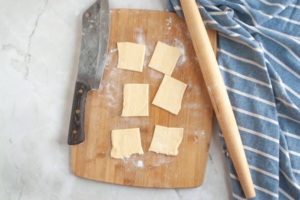 Overhead shot of square pieces of pale dough being cut on a wooden board with a knife and rolling pin.
