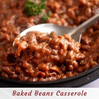 A close-up, slightly high-angle shot of a silver spoon scooping a portion of rich, reddish-brown baked beans casserole with visible pieces of meat and vegetables, garnished with fresh green parsley.