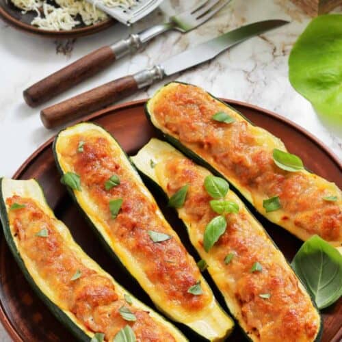 Four baked zucchini boats stuffed with a cheesy filling, garnished with fresh basil, served on a brown plate. Grated cheese and utensils are in the background.