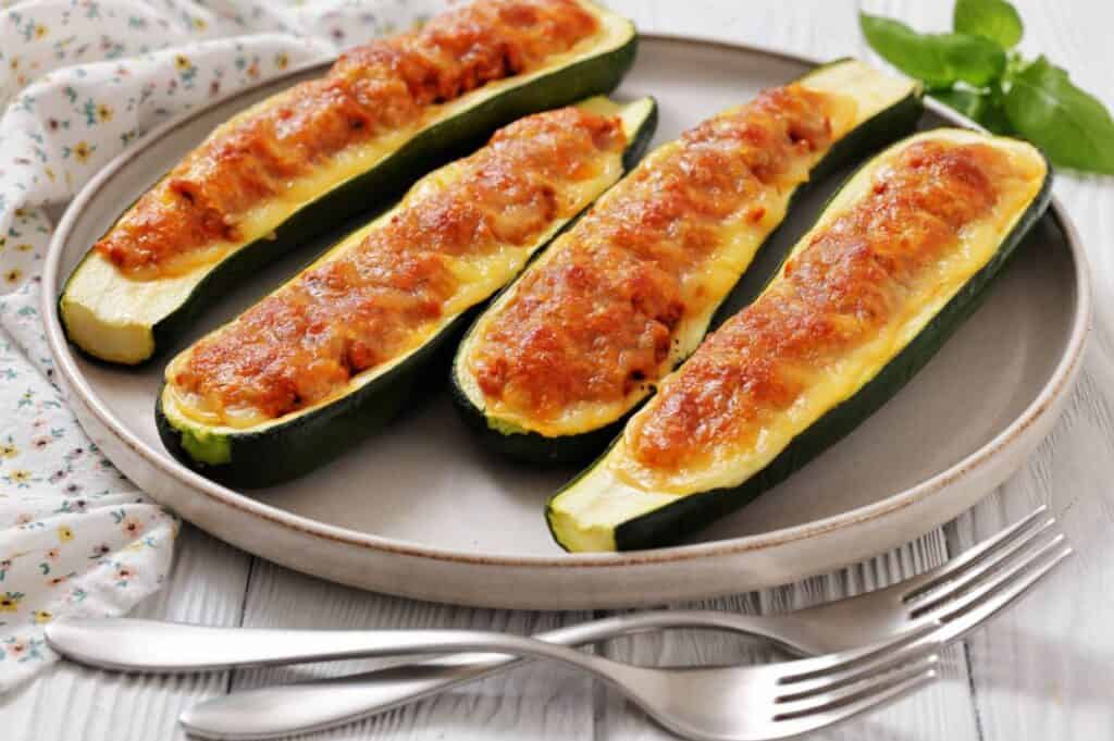 Four baked zucchini boats stuffed with a golden-brown cheese and tomato filling are arranged on a round plate with two forks beside it.