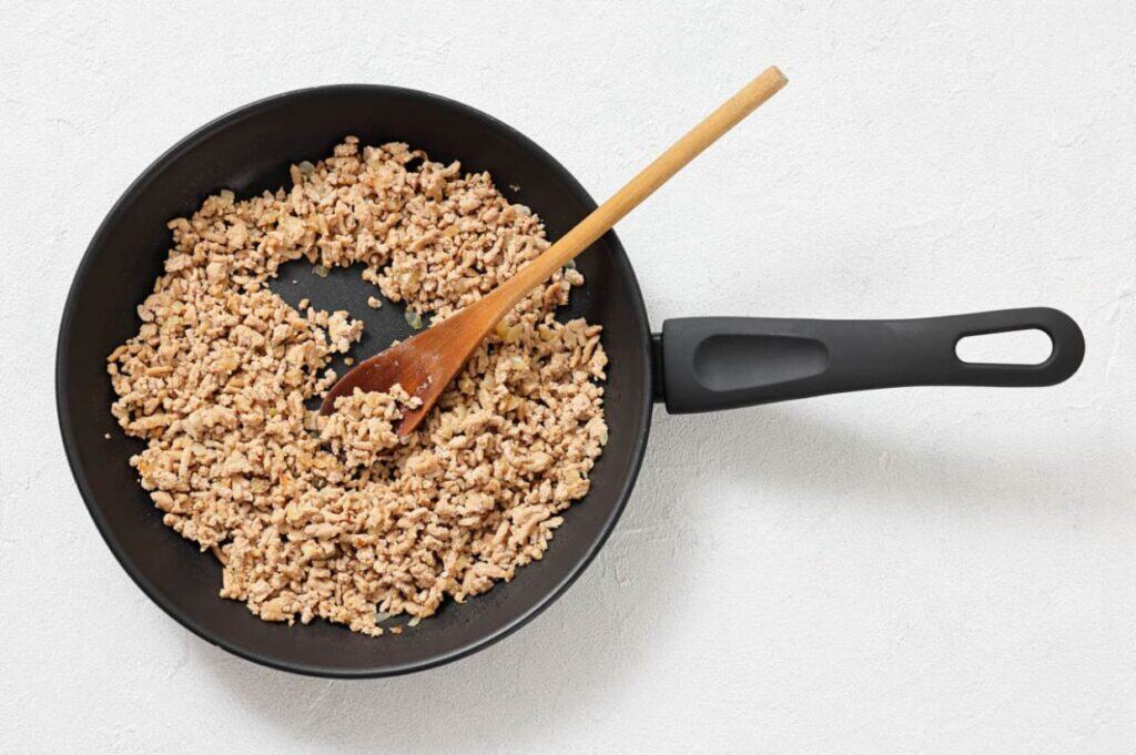 A frying pan with cooked ground meat being stirred by a wooden spoon, set on a white surface.