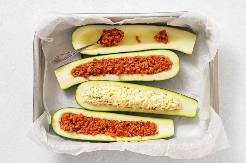 Three halved zucchinis in a baking dish, two filled with a tomato-based mixture, one topped with shredded cheese, and a spoon resting on one zucchini.