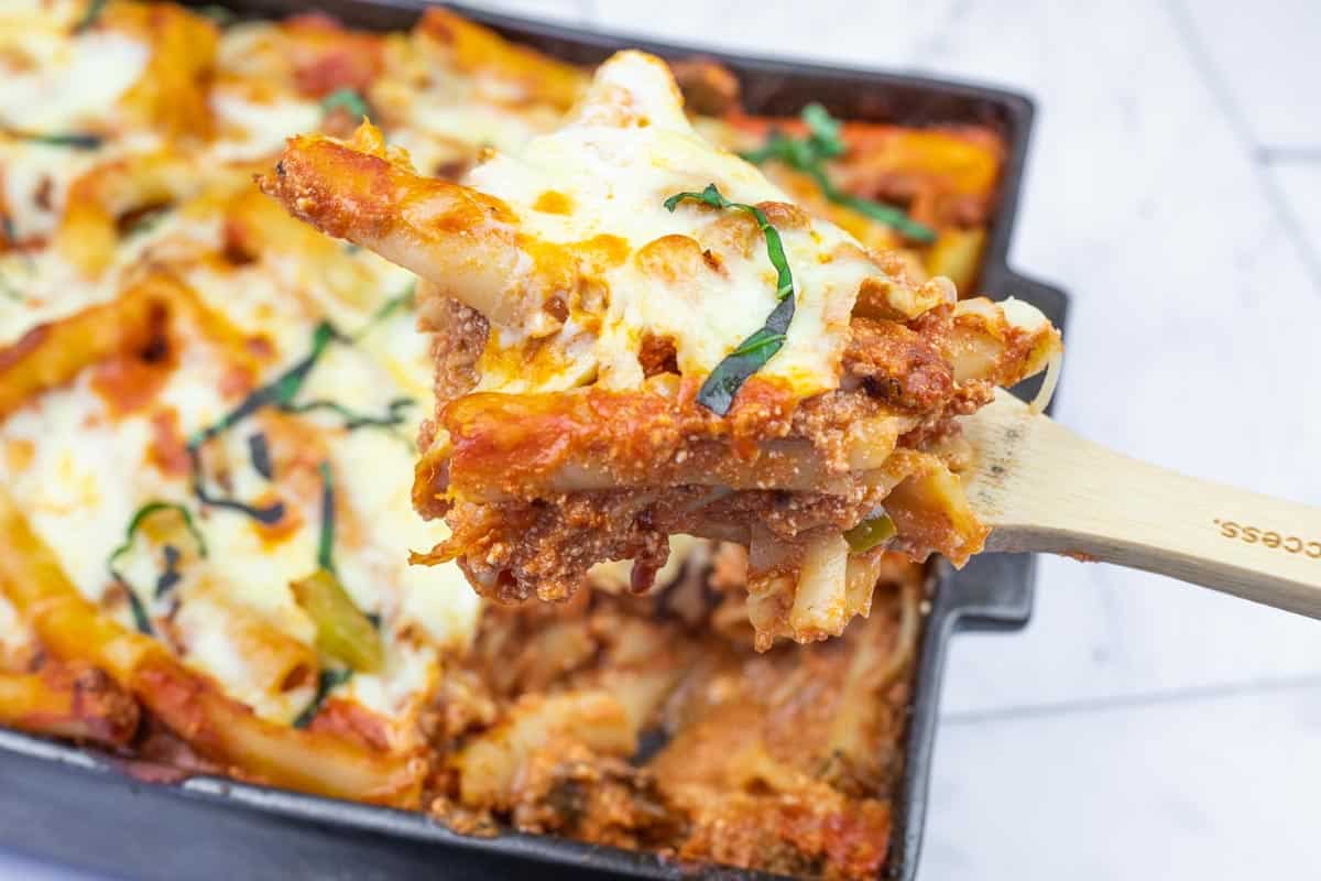 A close-up of a wooden spoon lifting a serving of baked ziti with melted cheese and tomato sauce from a casserole dish.