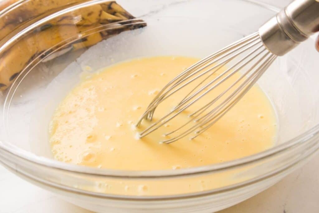 A metal whisk stirring a bowl of beaten eggs, with ripe bananas visible in the background.