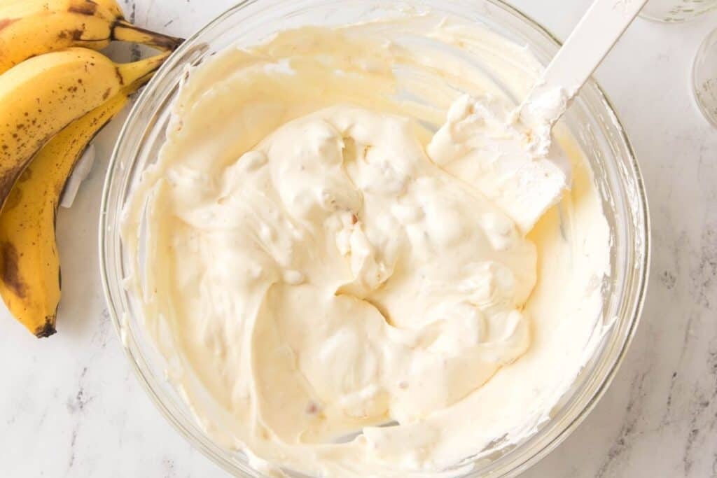 A glass bowl filled with creamy banana pudding mixture next to two whole bananas on a white marble surface.