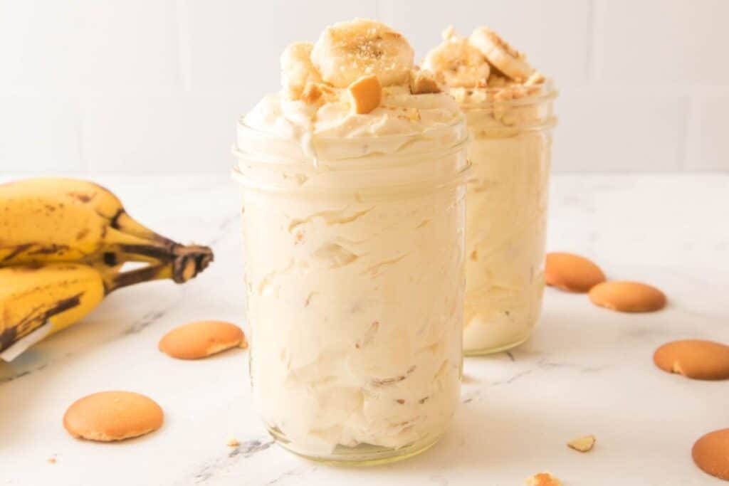 Two jars filled with banana pudding topped with banana slices and vanilla wafers, with whole bananas and vanilla wafers scattered on a white surface.
