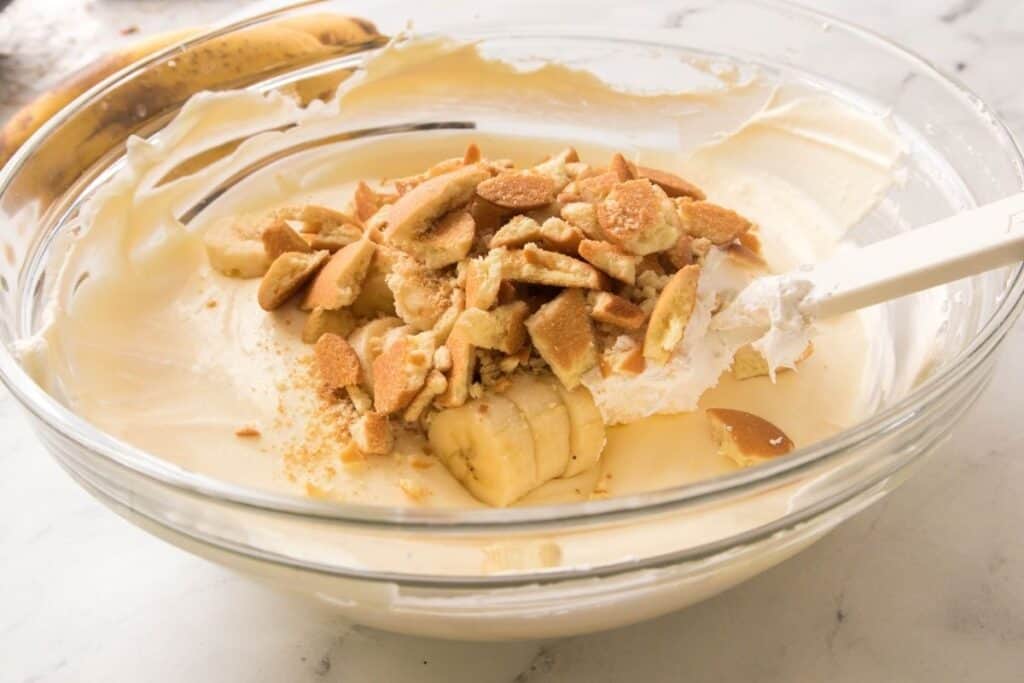 A glass bowl contains a creamy mixture with sliced bananas and crushed vanilla wafers on top, being stirred with a white spatula.