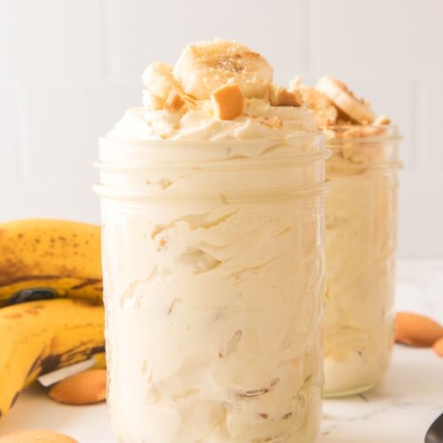 Two clear mason jars filled with creamy banana pudding, topped with sliced bananas and crushed vanilla wafers, with whole bananas and wafers in the background.