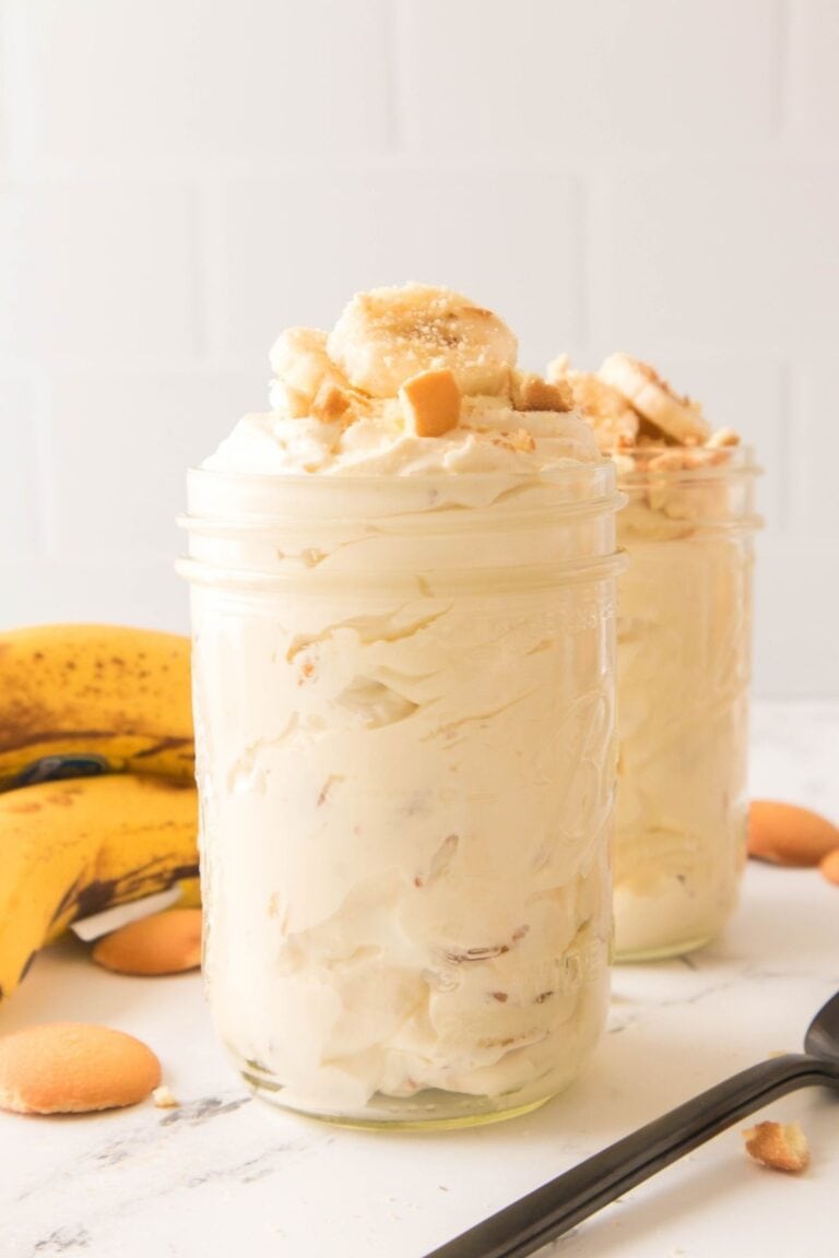 Two clear mason jars filled with creamy banana pudding, topped with sliced bananas and crushed vanilla wafers, with whole bananas and wafers in the background.