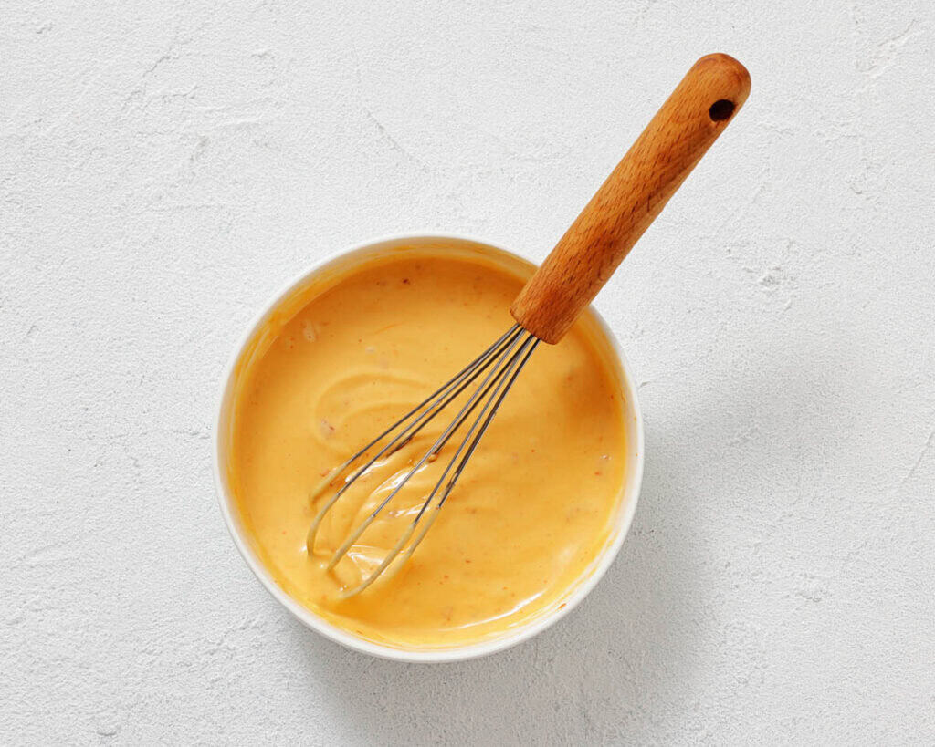 A white bowl filled with creamy orange sauce and a metal whisk with a wooden handle on a light textured surface.
