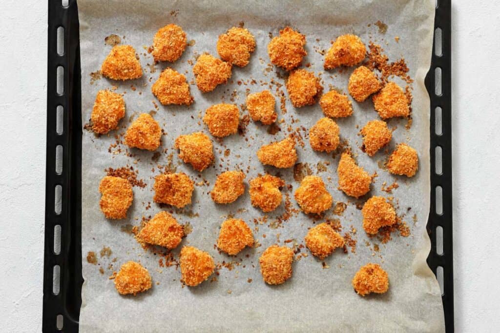 Baked, breaded cauliflower florets are spread out on a parchment-lined baking tray.