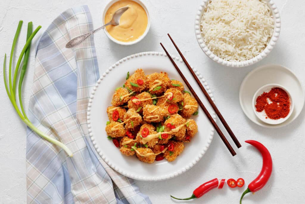 A plate of fried tofu garnished with red chili and green onions, served with a bowl of white rice, dipping sauce, chili powder, and whole chili peppers on a white surface.