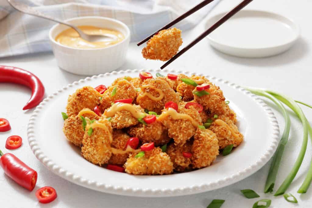 Plate of breaded, baked chicken pieces topped with sliced red chili, green onions, and a drizzle of sauce; chopsticks holding one piece; dipping sauce and chili peppers nearby.