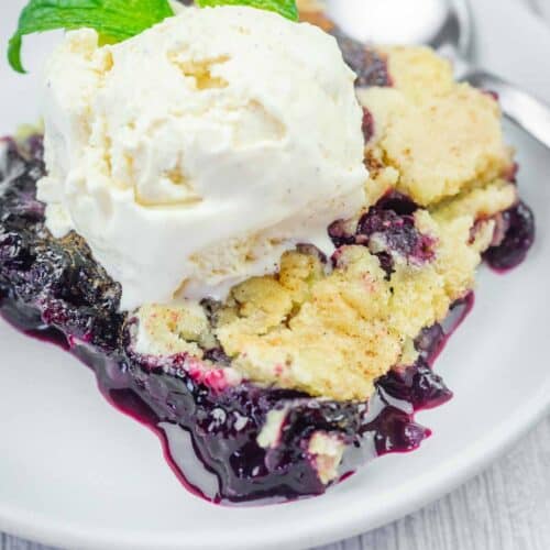 A serving of blueberry crumble topped with a scoop of vanilla ice cream and a sprig of mint on a white plate, with a spoon in the background.