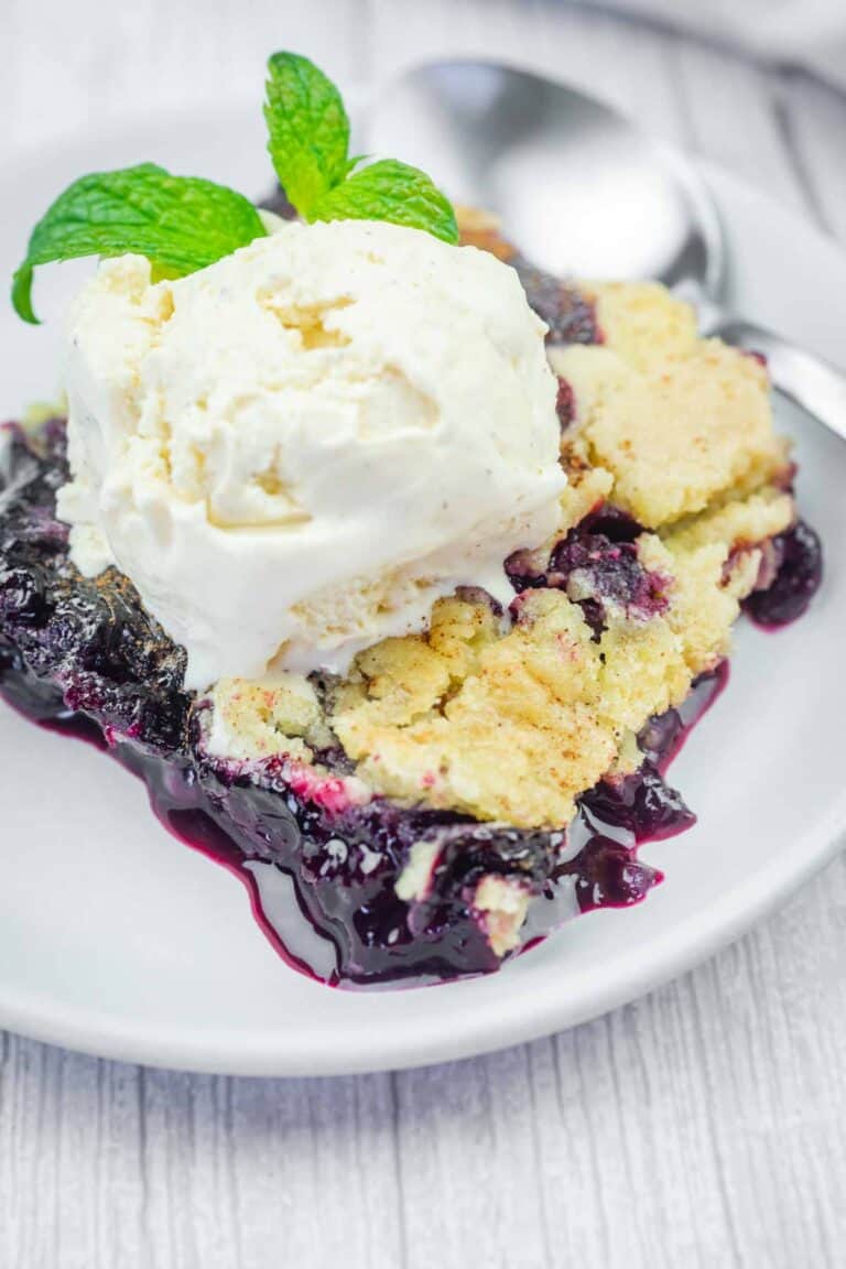 A serving of blueberry crumble topped with a scoop of vanilla ice cream and a sprig of mint on a white plate, with a spoon in the background.