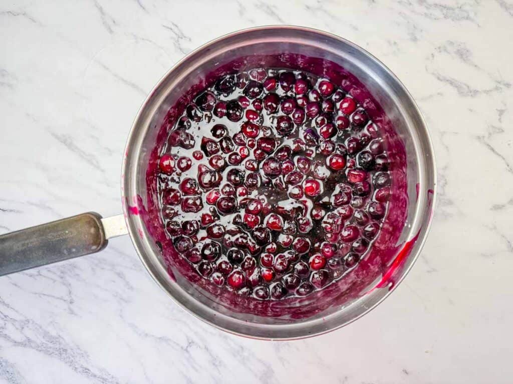 A saucepan filled with simmering blueberry mixture on a white marble countertop.