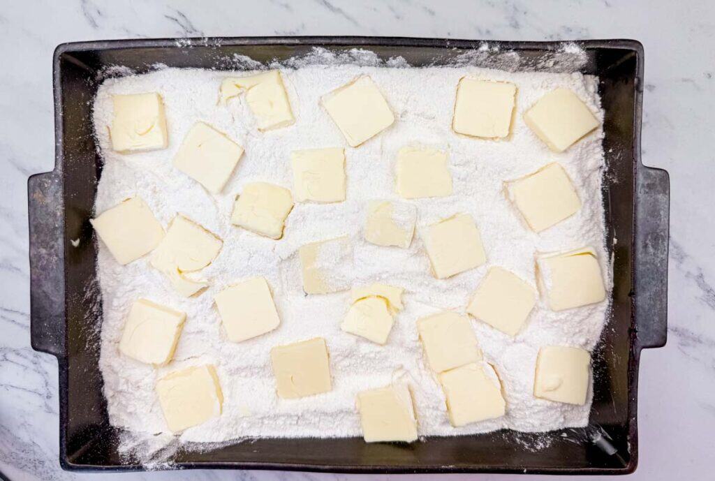 Rectangular baking pan filled with flour and topped with evenly spaced slices of butter, placed on a marble surface.