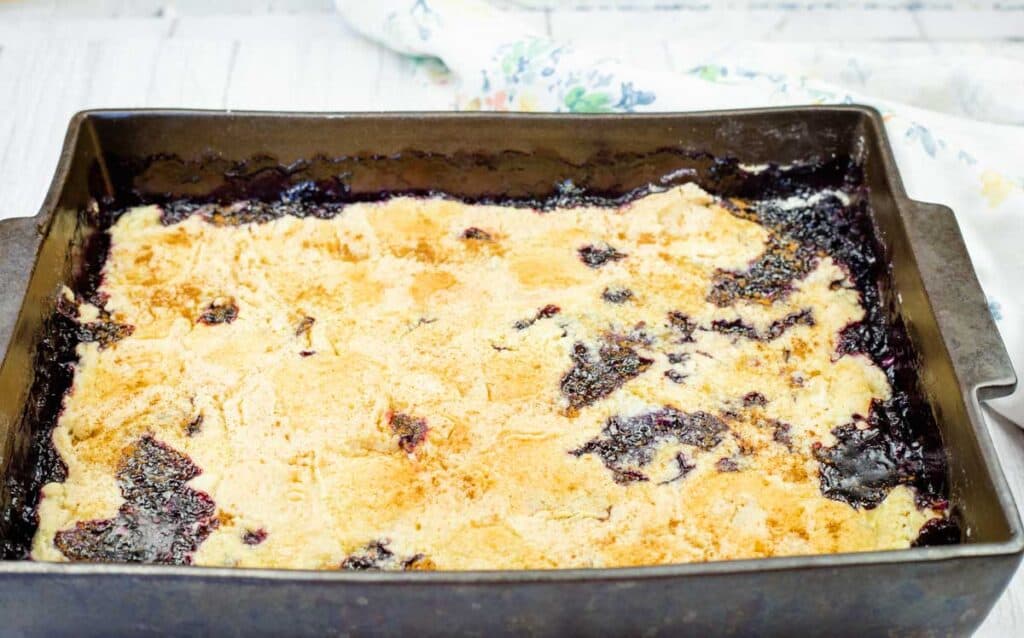 A rectangular metal baking dish filled with baked blackberry cobbler, featuring a golden-brown crust with patches of dark blackberry filling visible.