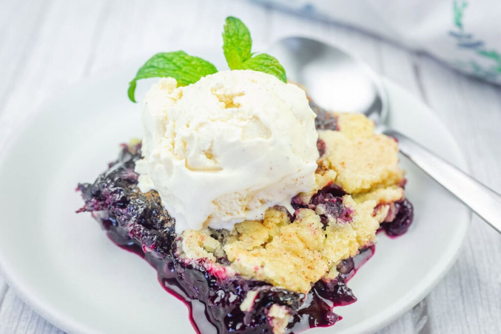 A serving of blueberry cobbler topped with a scoop of vanilla ice cream and a mint sprig, on a white plate with a spoon.