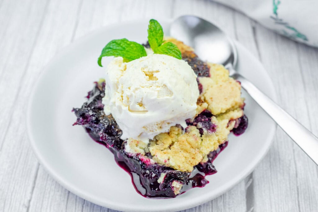 A serving of blueberry cobbler topped with a scoop of vanilla ice cream and a mint sprig, on a white plate with a spoon.