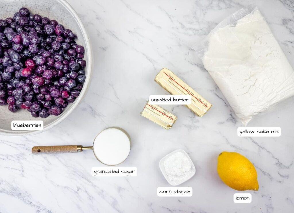 Ingredients for a blueberry dessert arranged on a marble surface: frozen blueberries, granulated sugar, cornstarch, unsalted butter, yellow cake mix, and a lemon.
