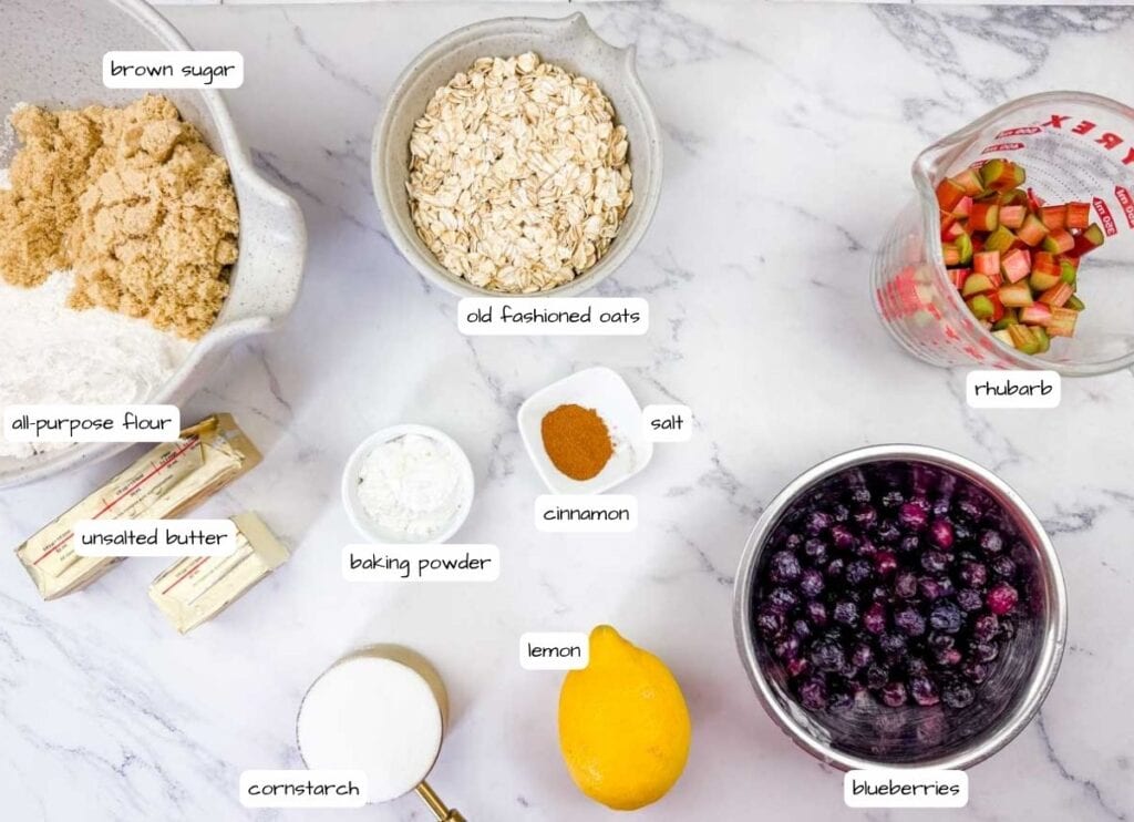 Overhead shot of labeled ingredients for Blueberry Rhubarb Bars: brown sugar, all-purpose flour, unsalted butter, old-fashioned oats, baking powder, cinnamon, salt, cornstarch, lemon, rhubarb, and blueberries.