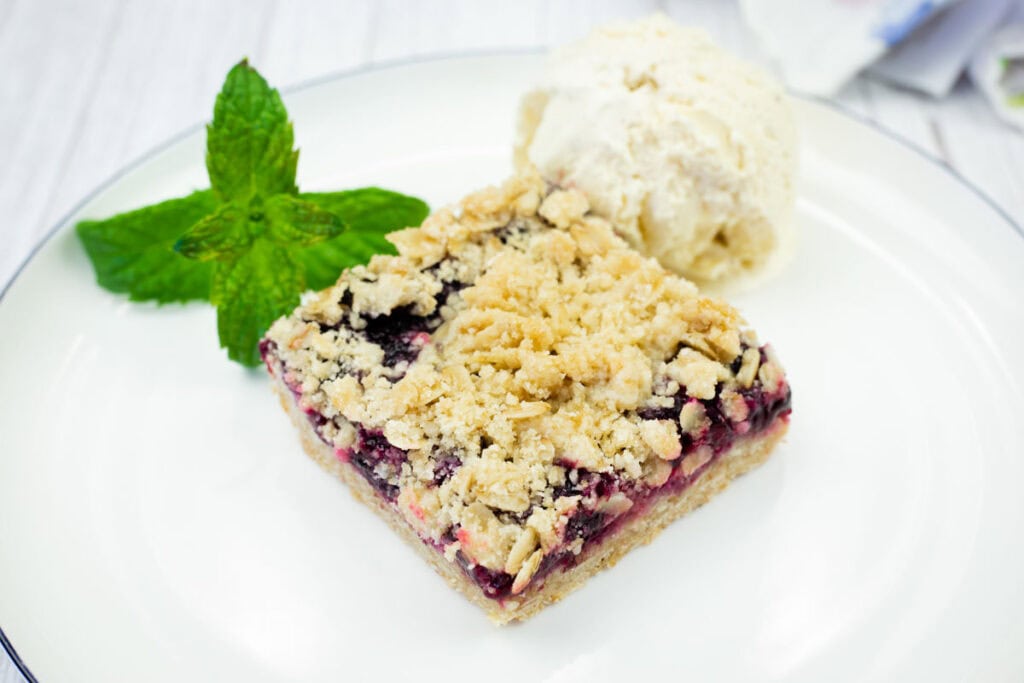 A white plate holds a square Blueberry-Rhubarb Crumble Bar topped with a scoop of vanilla ice cream and fresh mint, with a colorful cloth in the background.
