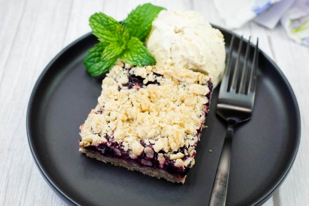 A black plate holds a square Blueberry-Rhubarb Crumble Bar topped with a scoop of vanilla ice cream and fresh mint, with a fork on the side.