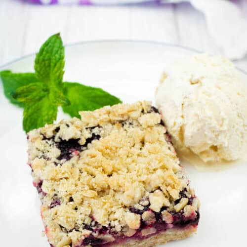 A white plate holds a square Blueberry-Rhubarb Crumble Bar topped with a scoop of vanilla ice cream and fresh mint, with a colorful cloth in the background.