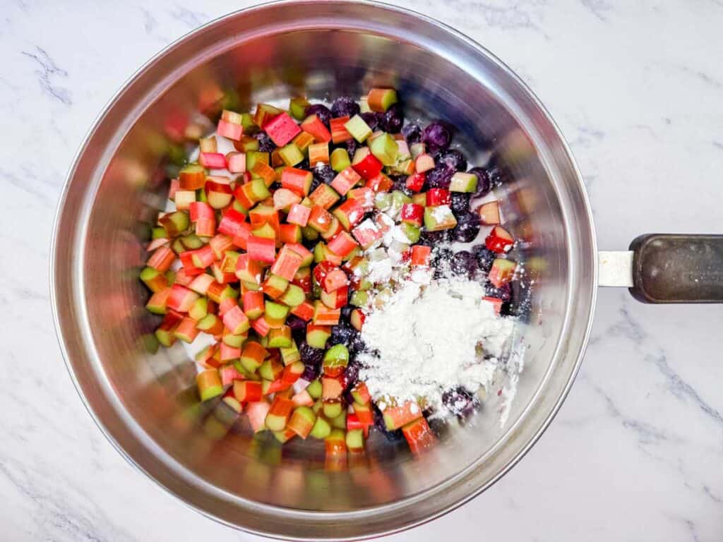 Overhead shot of a stainless steel saucepan filled with fresh chopped red and green rhubarb, dark blue blueberries, and a pile of white cornstarch, ready to be cooked.