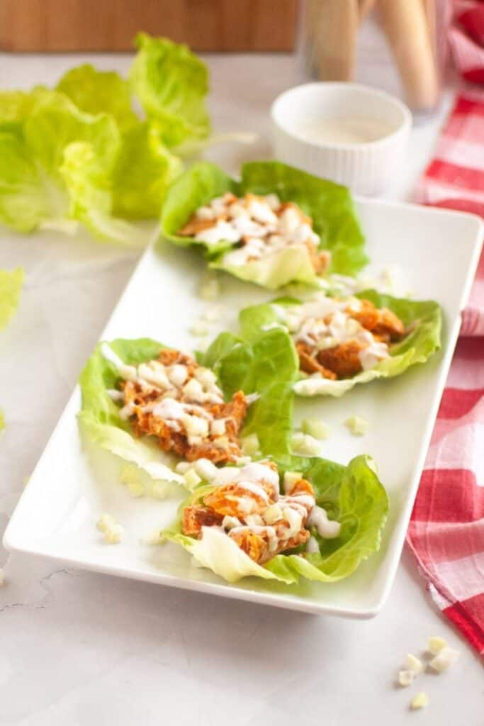 Four lettuce wraps filled with shredded chicken, chopped onions, and drizzled with a white sauce are arranged on a rectangular white plate beside a red checkered napkin.