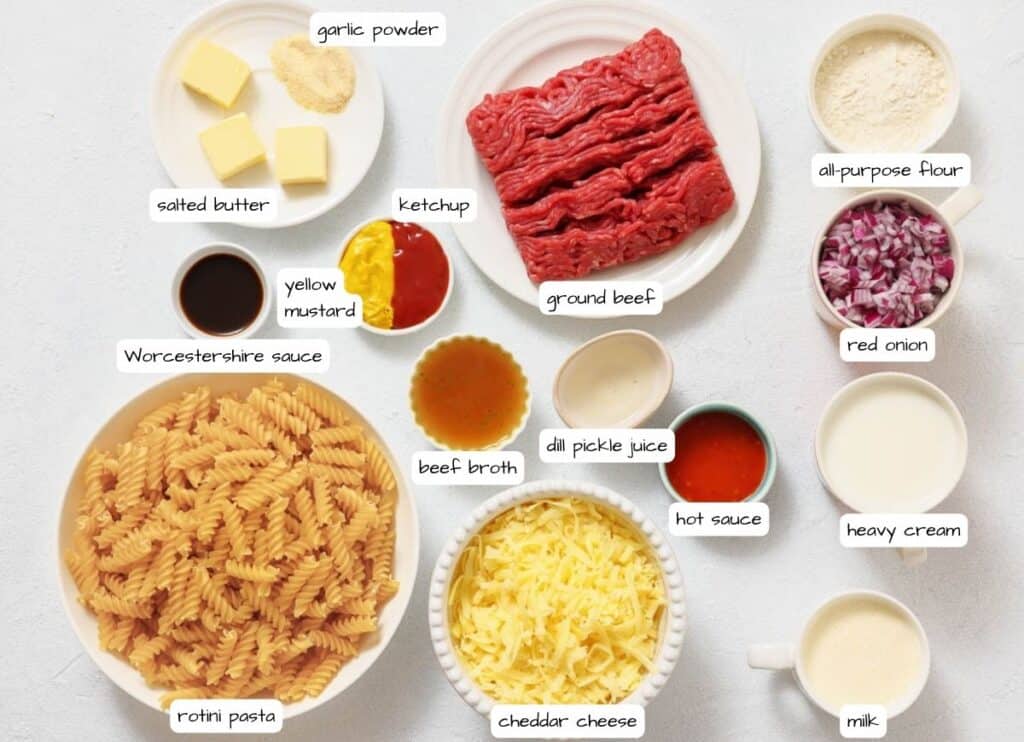Ingredients for a beef pasta dish are arranged on a white surface, each labeled, including ground beef, rotini pasta, cheddar cheese, and various sauces, spices, and dairy products.