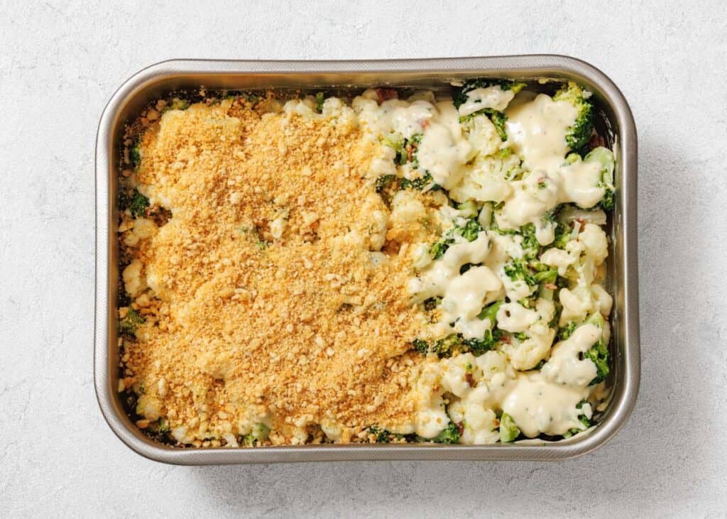 A metal baking dish containing a broccoli and cauliflower casserole, half topped with breadcrumbs and half with cheese sauce, on a light surface.