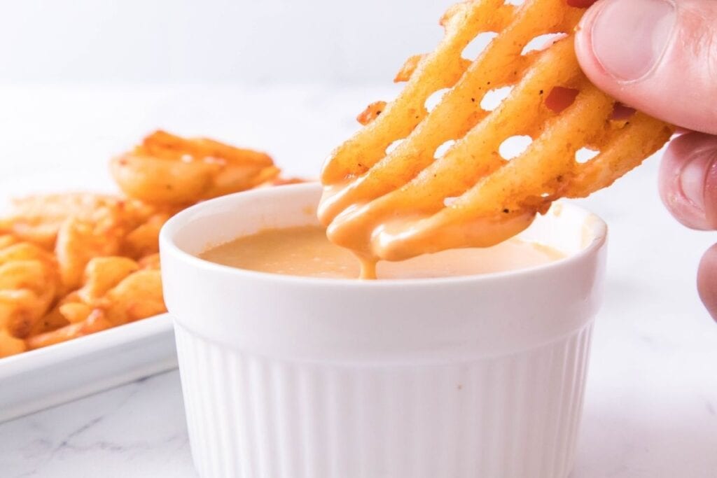 A hand dips a waffle fry into a small white ramekin of creamy dipping sauce, with more fries visible in the background.