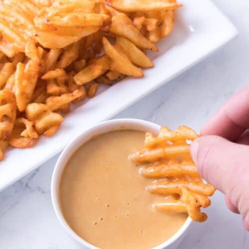 A hand dips a seasoned waffle fry into a small bowl of creamy sauce, with more fries on a rectangular white plate in the background.