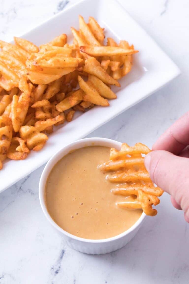 A hand dips a seasoned waffle fry into a small bowl of creamy sauce, with more fries on a rectangular white plate in the background.