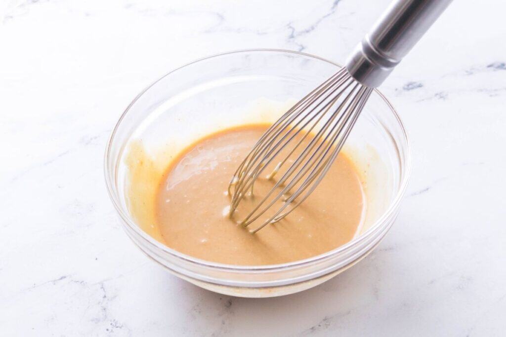 A metal whisk rests in a clear glass bowl containing a smooth, tan-colored batter on a white marble surface.