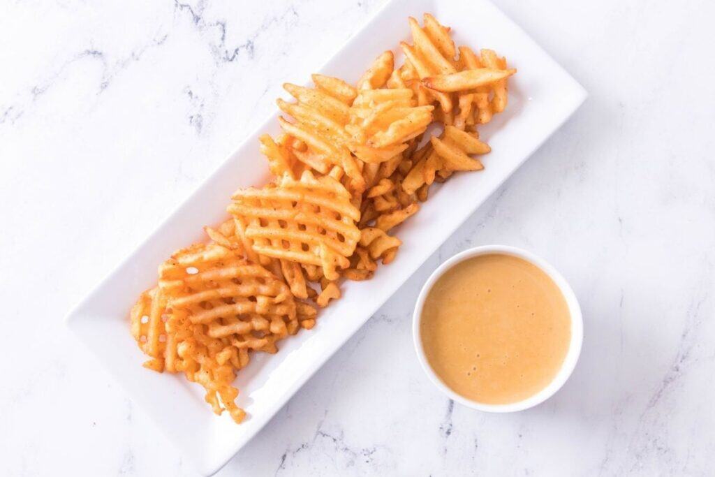 Waffle fries arranged on a rectangular white plate next to a small cup of dipping sauce on a white marble surface.