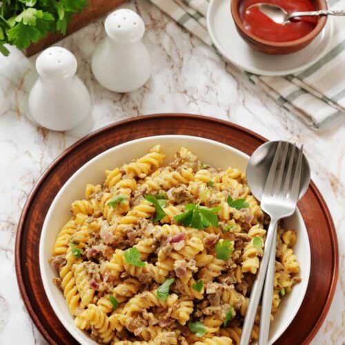 A bowl of cooked fusilli pasta mixed with ground meat and topped with parsley, placed on a wooden plate with a fork and spoon. Salt and pepper shakers, ketchup, and greens are in the background.
