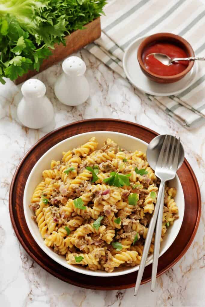 A bowl of cooked fusilli pasta mixed with ground meat and topped with parsley, placed on a wooden plate with a fork and spoon. Salt and pepper shakers, ketchup, and greens are in the background.