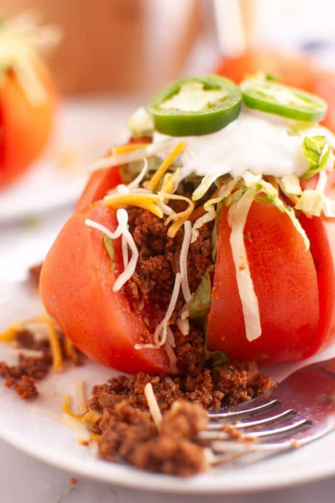 A close-up of a red tomato hollowed out and stuffed with ground taco meat, shredded lettuce, cheese, sour cream, and sliced jalapenos, on a white plate with a fork.