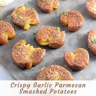 Overhead shot of several golden-brown Crispy Garlic Parmesan Smashed Potatoes on a rustic metal baking sheet, garnished with fresh parsley.
